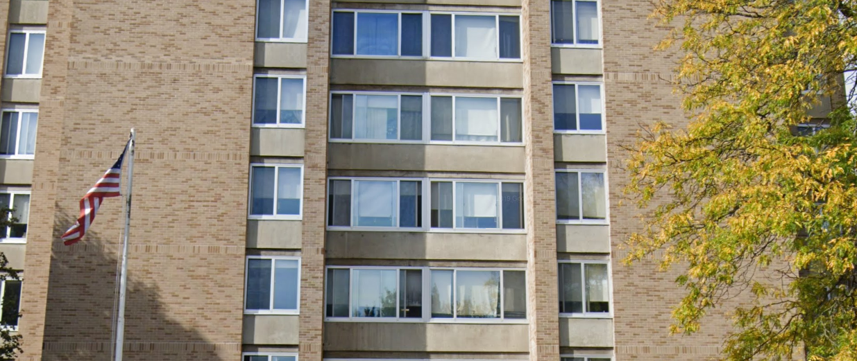 A mid-rise brick building with large windows, an American flag on a flagpole in front, and a tree with yellow leaves on the right side.