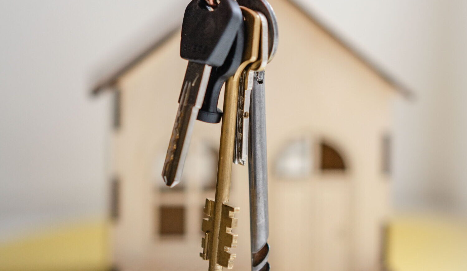 A set of house keys on a keyring is in focus, with a small wooden model of a house blurred in the background.