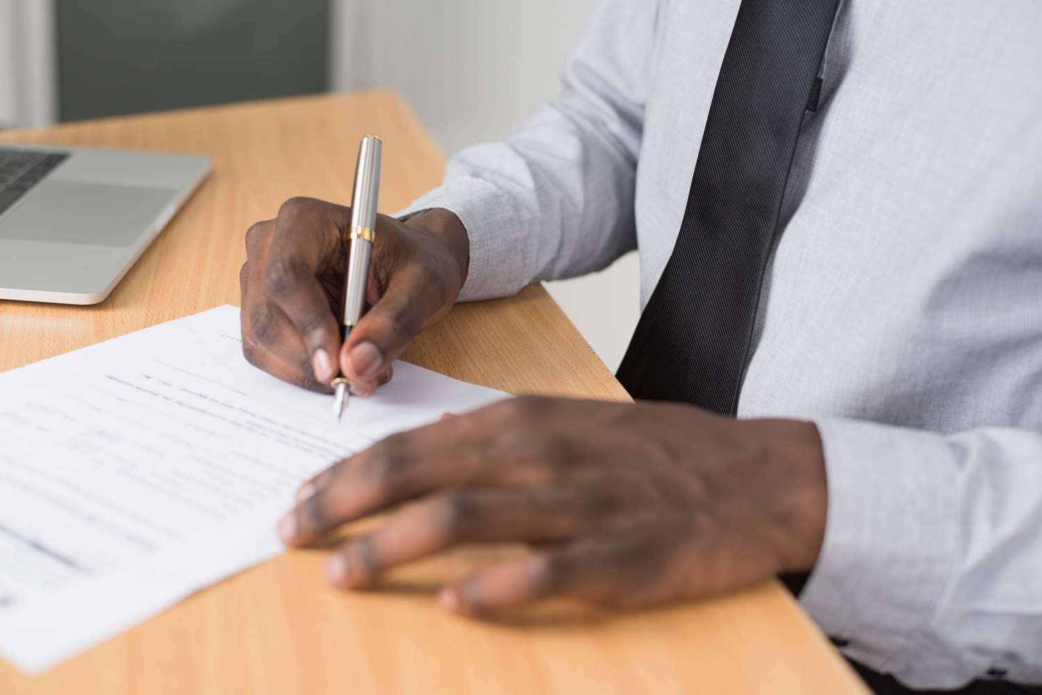 A person in a shirt and tie sits at a desk, holding a pen and signing a document, with a laptop partly visible beside them.